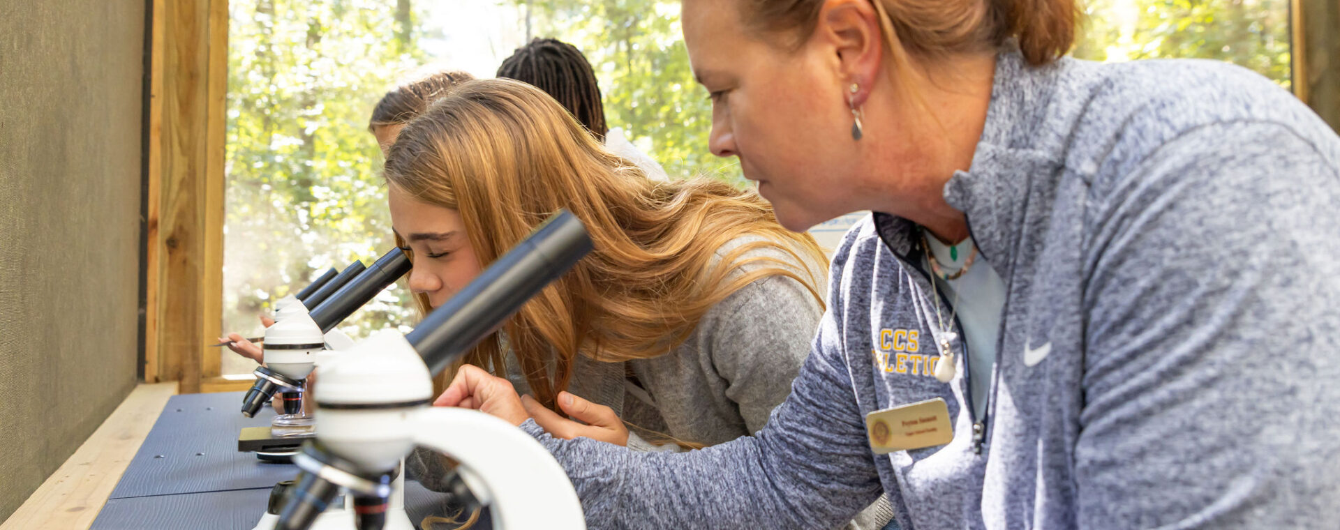 A CCS upper school teacher looks through a microscope with students in the outdoor education center. 