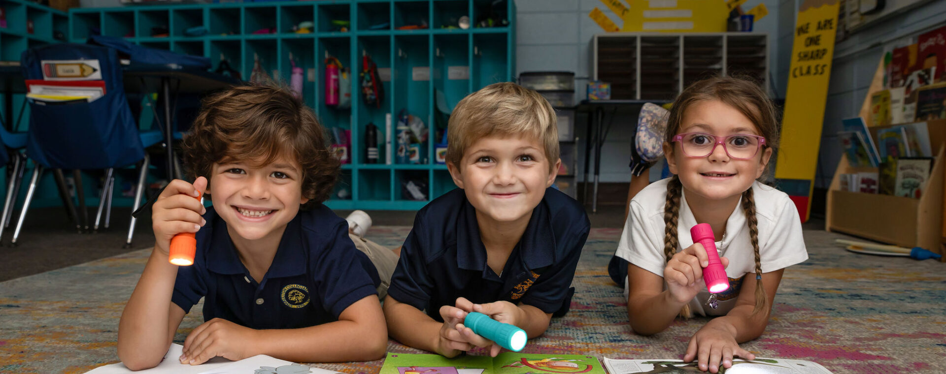 Three young students laying on the classroom floor reading books.
