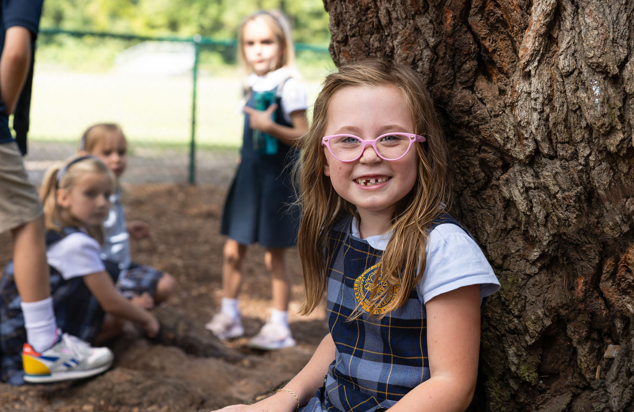 A smiling lower school student with their classmates outside under a tree.
